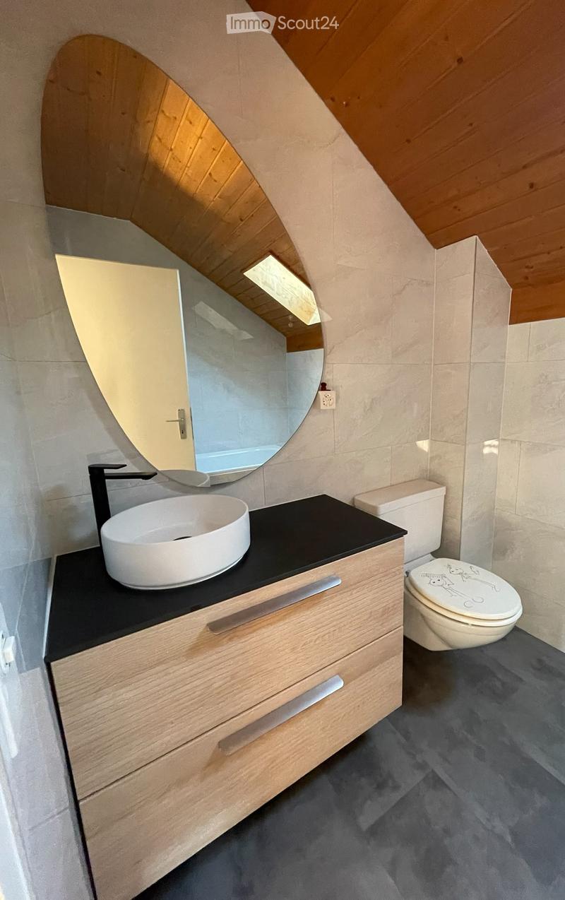 Modern bathroom with a sleek wooden vanity, black countertop, white sink, and a round mirror above it. A toilet is positioned next to the vanity, and the room features a sloped wooden ceiling and grey tiled walls.