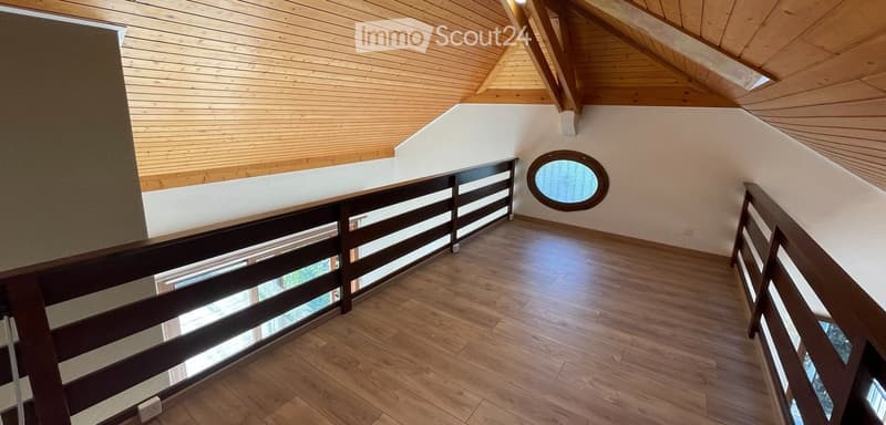 Spacious hallway with wooden flooring, slanted wooden ceiling, and dark wooden railing featuring glass panels. A round window is visible on the wall.