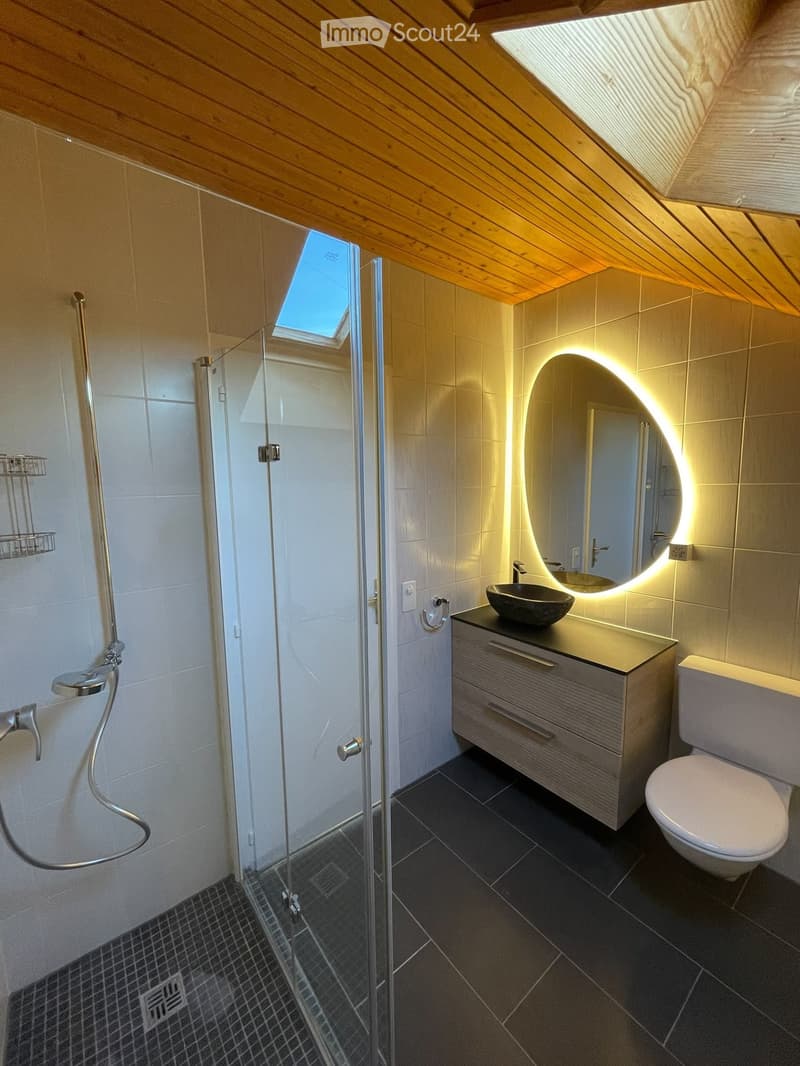 Modern bathroom with a glass-enclosed shower, a sleek sink with a modern faucet, a round illuminated mirror, and a toilet. The space features dark tiled floors and light-colored walls.