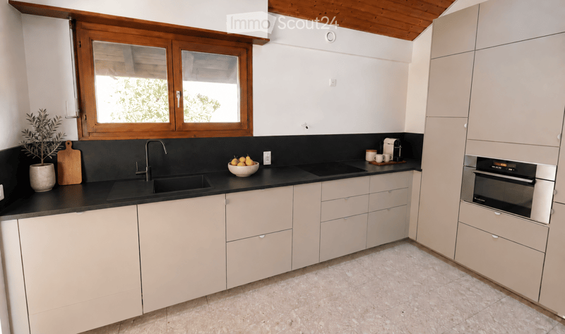 Modern kitchen with light-colored cabinets, black countertops, a sink, and an oven. Features a window with wooden frames, a potted plant, and a bowl of fruit on the counter.