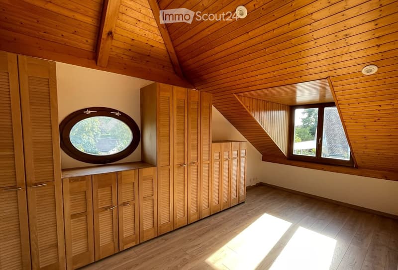 Bright, empty attic room with wooden walls, ceiling, and flooring. Features built-in wooden cabinets, a round window, and a large window with a view of the outside.