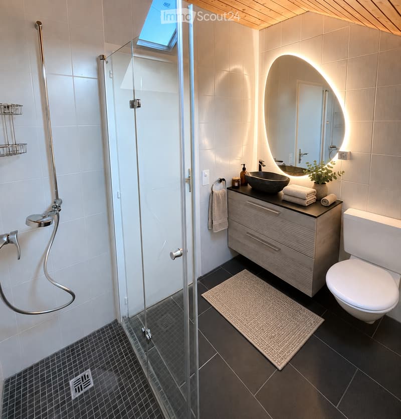 Modern bathroom with a glass-enclosed shower, a sleek sink with a modern mirror, a toilet, and a small plant on the counter. The space features a skylight and a warm, ambient light around the mirror.