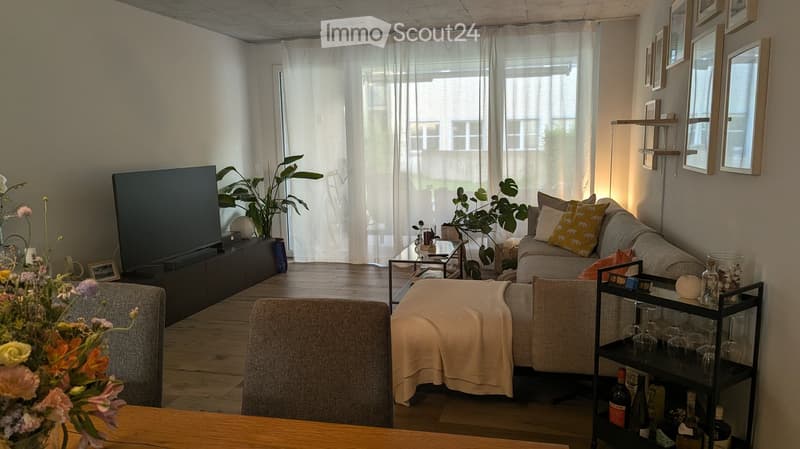 Modern living room with a grey sectional sofa, coffee table, ottoman, large television, and decorative plants. The space features large windows with sheer curtains and a wooden floor.