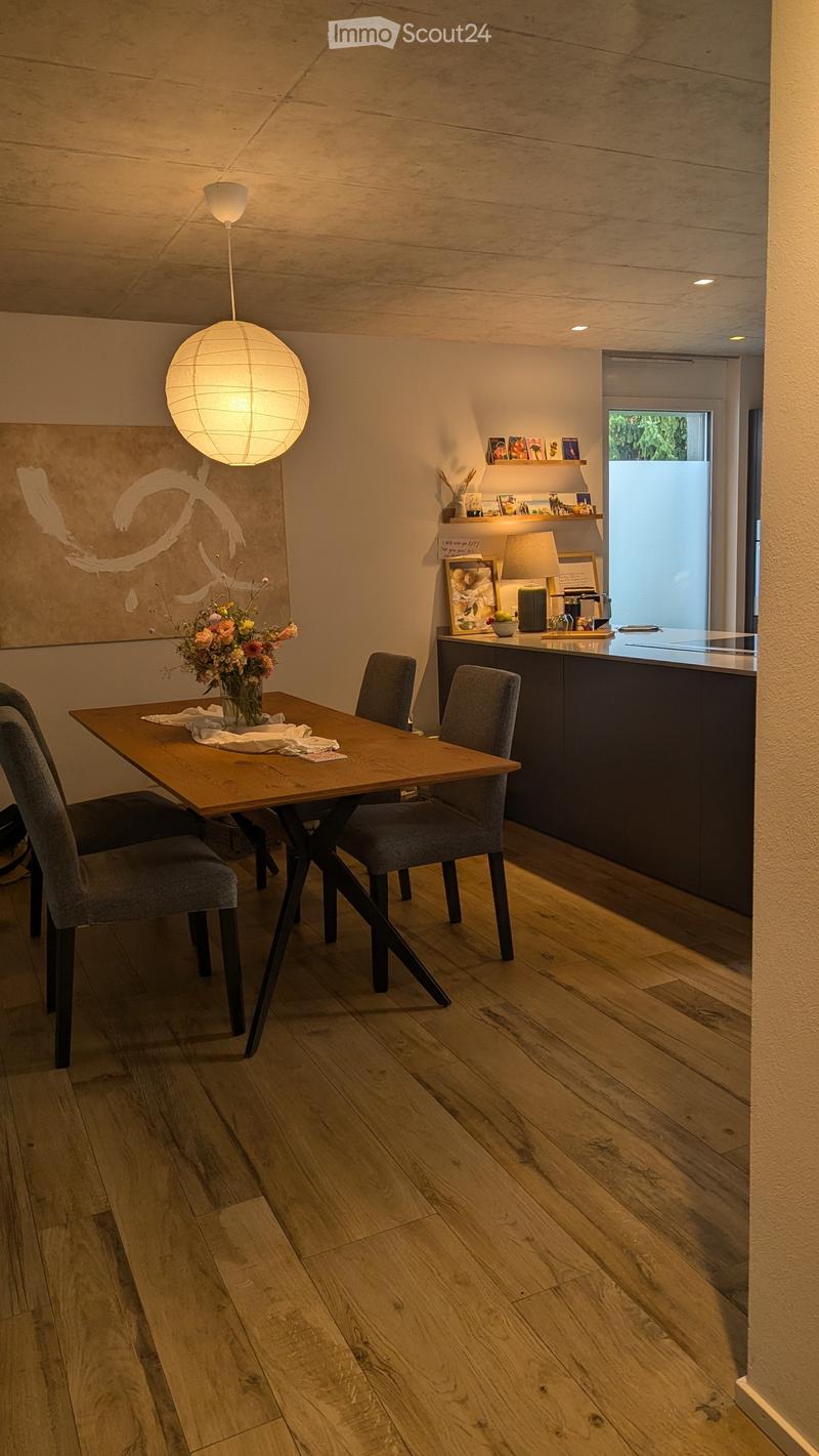 Modern kitchen and dining area with wooden flooring, a round pendant light, a wooden dining table with four chairs, and a vase of flowers on the table. The kitchen features a dark countertop with various items, including a lamp and framed pictures.