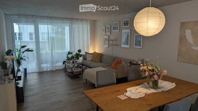 Modern living room with wooden flooring, large window with sheer curtains, grey sofa with colorful cushions, coffee table, dining table with chairs, and decorative items including a large pendant light and artwork on the walls.