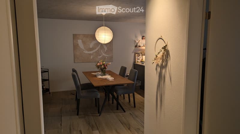 Modern dining area with a wooden table, four grey upholstered chairs, a vase with flowers, a round pendant light, and a decorative mirror on the wall.