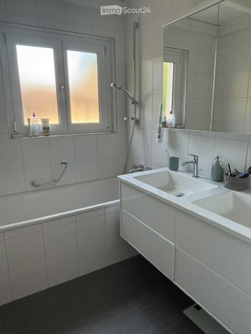 Modern bathroom with a bathtub, double sink, large mirror, and a window with frosted glass. Includes a handrail next to the bathtub and a showerhead above it.