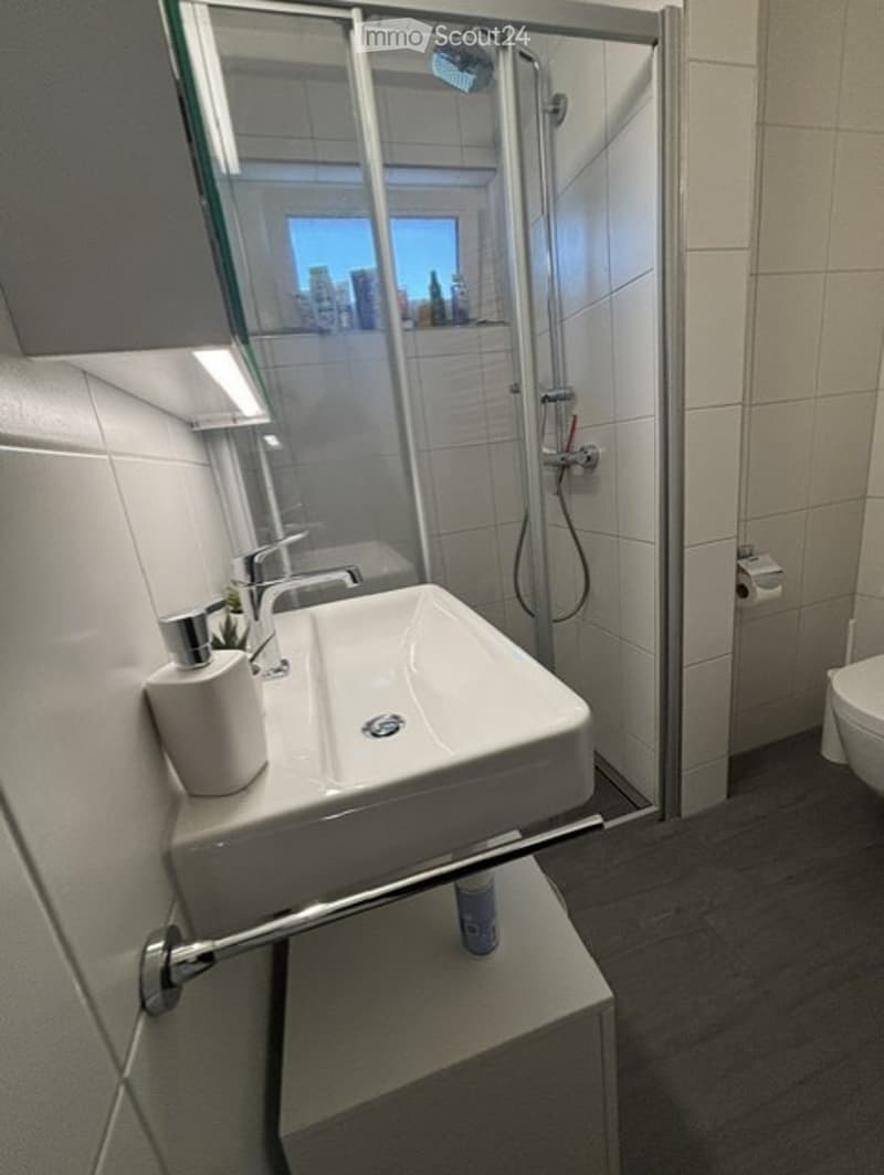 Modern bathroom with a white sink, chrome faucet, glass-enclosed shower, and a toilet. The space features white tiles and a dark grey floor.
