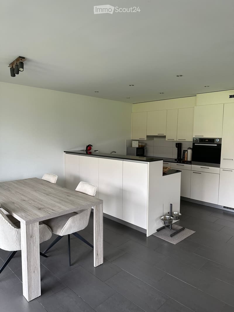 Modern kitchen with white cabinets, black countertops, a wooden dining table with four chairs, and various appliances including an oven, microwave, and coffee maker.