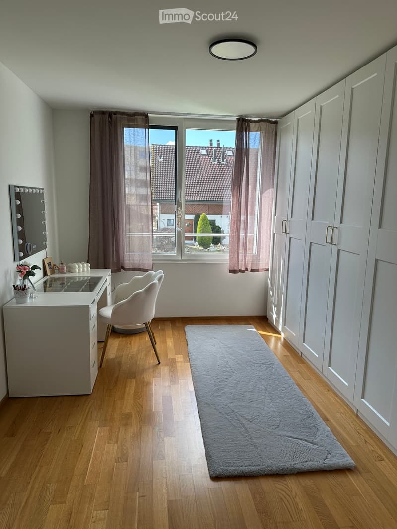 Well-lit room with wooden flooring, large window with view, white chair, vanity desk, mirror, and built-in wardrobes.
