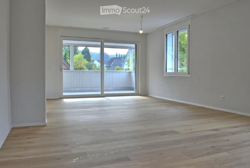 Empty room with wooden flooring, large sliding glass doors leading to a balcony, and a window on the right side.