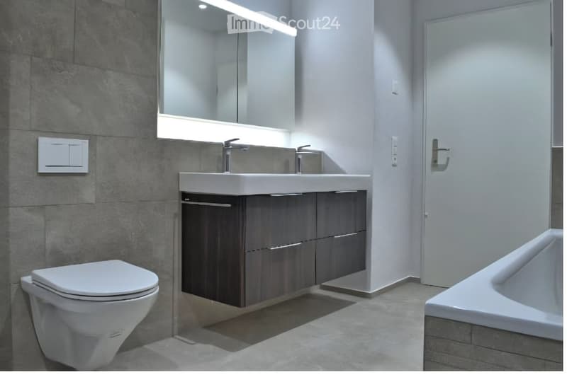 Modern bathroom with a white toilet, double sink vanity with dark wood cabinets, large mirror with built-in lighting, and a bathtub.