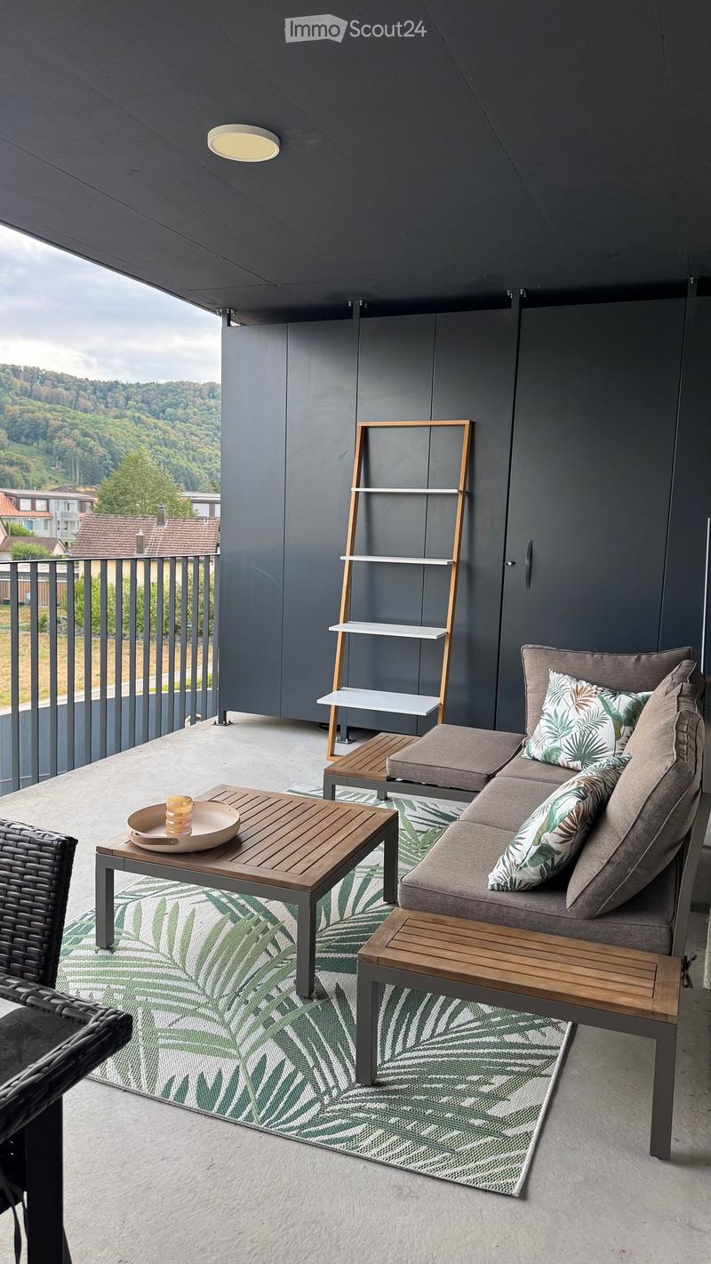 Modern balcony with grey sectional sofa, wooden coffee table, wicker chair, and a shelf unit, overlooking a green landscape.