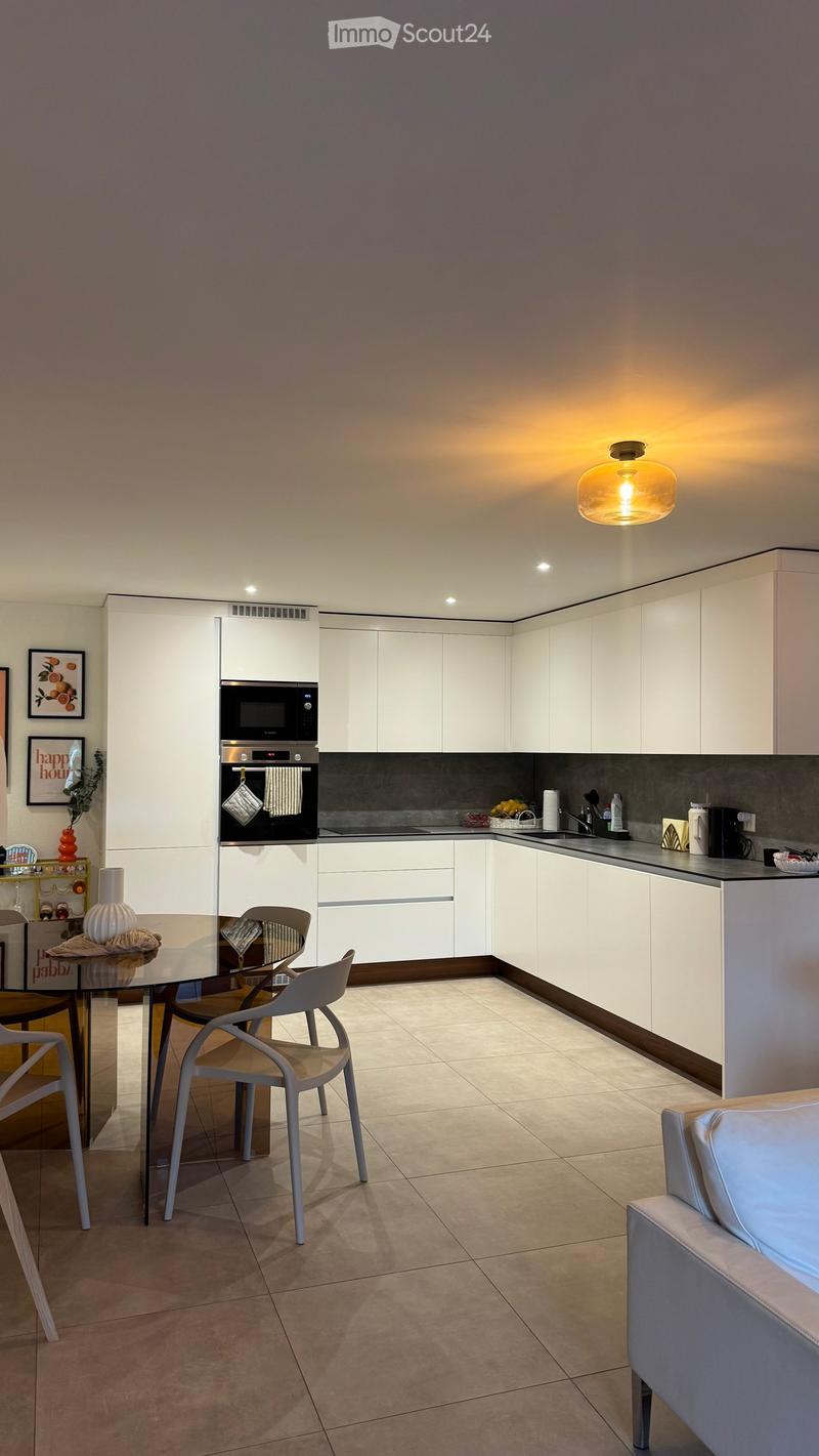 Modern kitchen with white cabinets, glass dining table, chairs, and various appliances including an oven, microwave, and coffee maker. The space features a sleek design with a tiled floor and a pendant light.