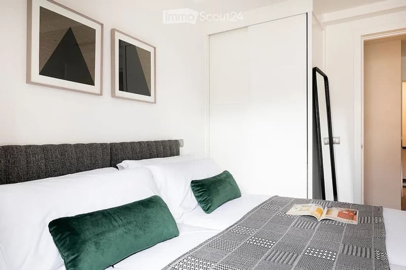 Modern bedroom with a large bed, grey headboard, white pillows, green throw pillows, grey and white patterned blanket, and two picture frames on the wall.