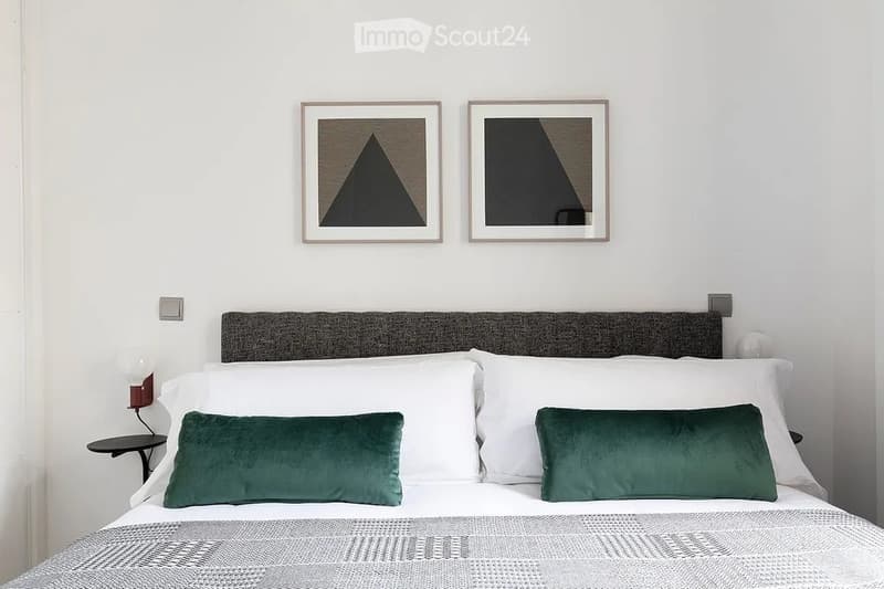 Modern bedroom with a neatly made bed, two green throw pillows, a nightstand with a lamp, and two framed abstract art pieces on the wall.