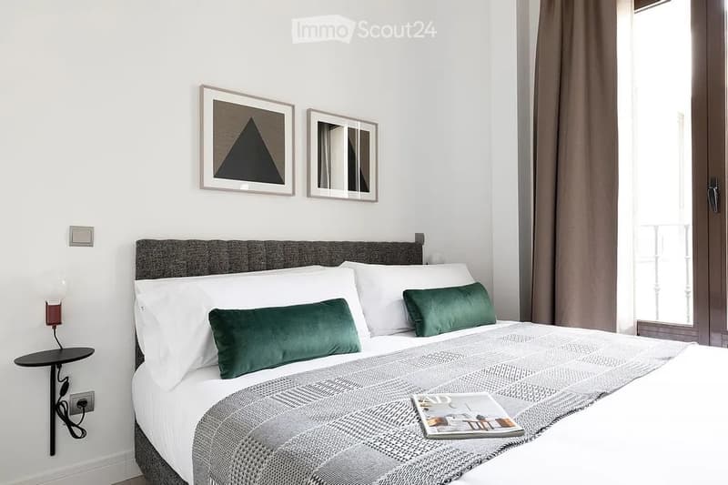 Modern bedroom with a neatly made bed, green accent pillows, a wall-mounted nightstand lamp, framed artwork, and a mirror above the bed.
