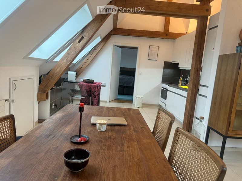 A modern, well-lit room with wooden beams and skylights, featuring a dining table with chairs, a laptop, and a kitchen area with white cabinets, a refrigerator, and an oven.
