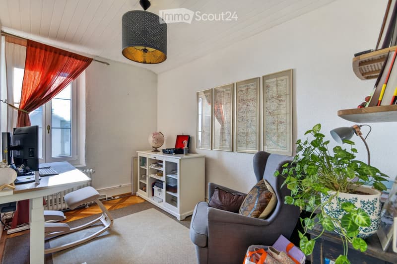 A cozy, well-furnished room with a grey sofa, armchair, white desk, computer, hanging lamp, plant, and decorative items including framed maps and a globe.