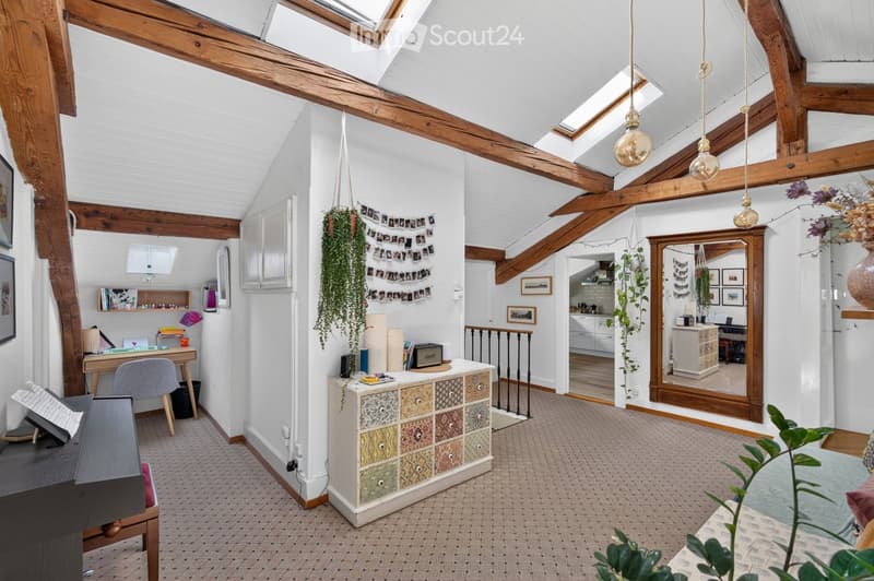 Cozy, loft-style room with exposed wooden beams, skylights, and modern decor. Features a desk with a chair, a small table with a mirror, a colorful cabinet, a piano, and several potted plants.