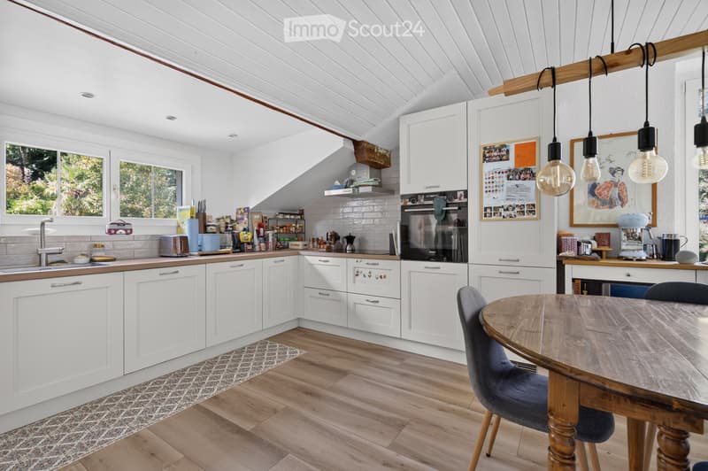 Bright, airy kitchen with white cabinets, wooden countertops, and a wooden dining table with chairs. Features include a sink, oven, toaster, coffee machine, and various kitchen items on the counter.