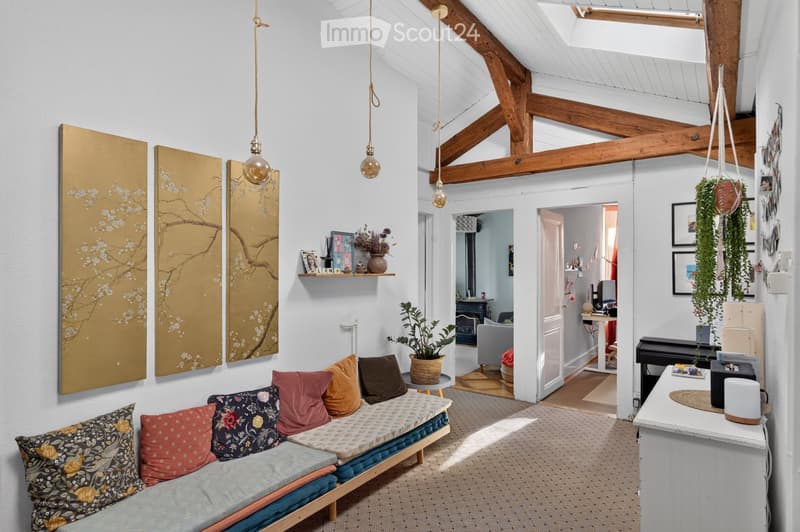 A cozy living space with a wooden ceiling, exposed beams, and hanging pendant lights. Features a daybed with colorful pillows, a small table with a potted plant, a shelf with decorative items, and a piano in the corner.