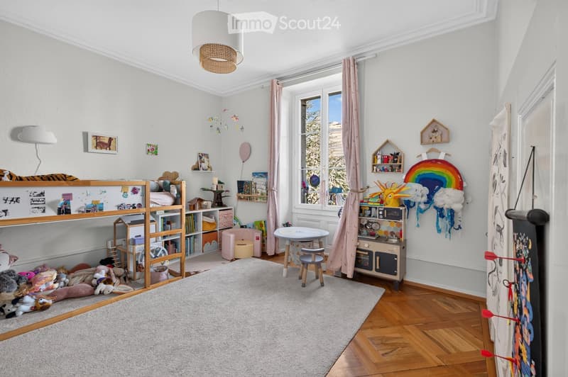 A well-lit children's bedroom with a bunk bed, shelves filled with books and toys, a small table and chair, and a large window with pink curtains. The room features a colorful toy rainbow and a cloud mobile hanging from the ceiling.