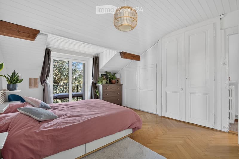 Cozy attic bedroom with wooden floors, white walls, slanted ceiling, and a view of trees through a large window. Features a double bed with pink bedding, a nightstand, a wicker pendant light, and a dresser with potted plants.