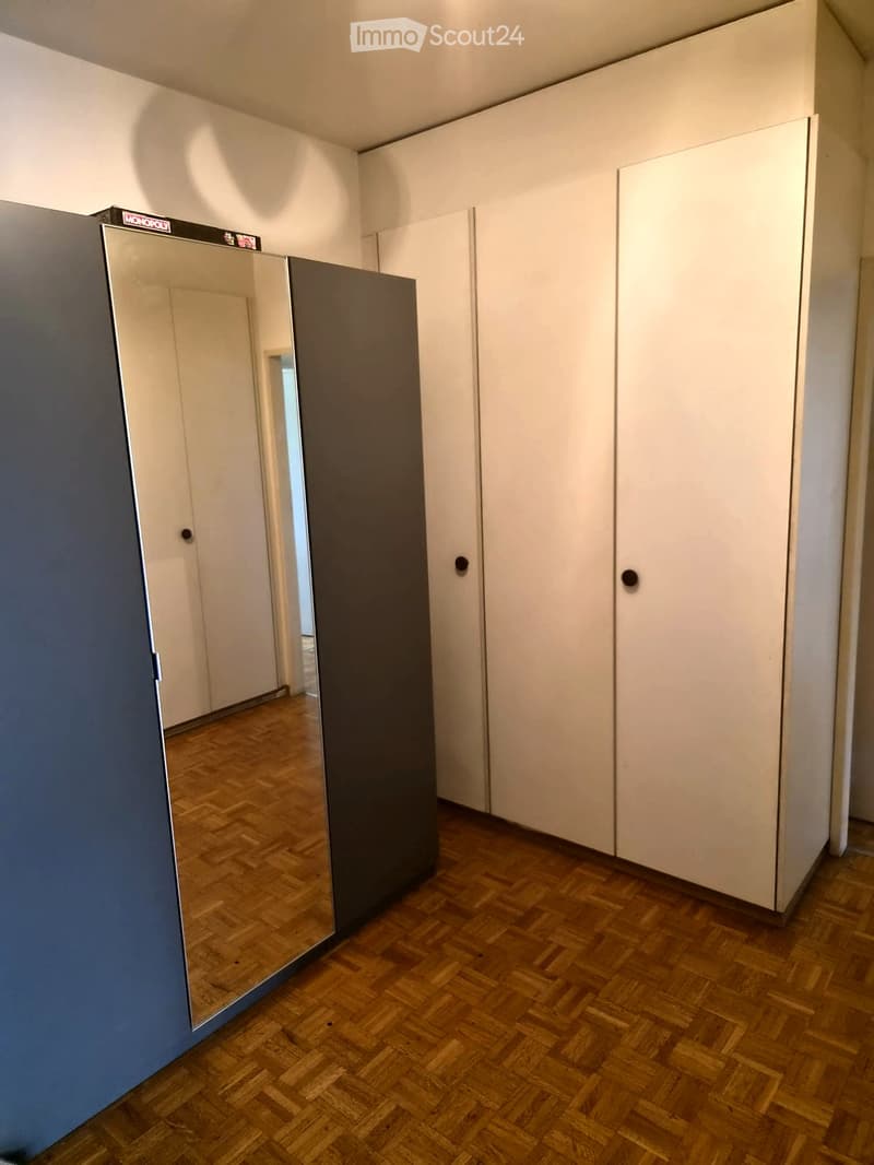 Empty room with wooden herringbone flooring, white built-in wardrobes, and a large mirror on the wall.