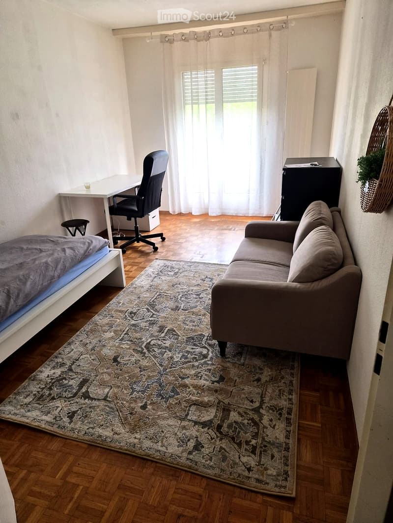 A compact, well-furnished room with a bed, sofa, desk, and chair. The room features wooden flooring, a patterned rug, and large windows with sheer curtains.
