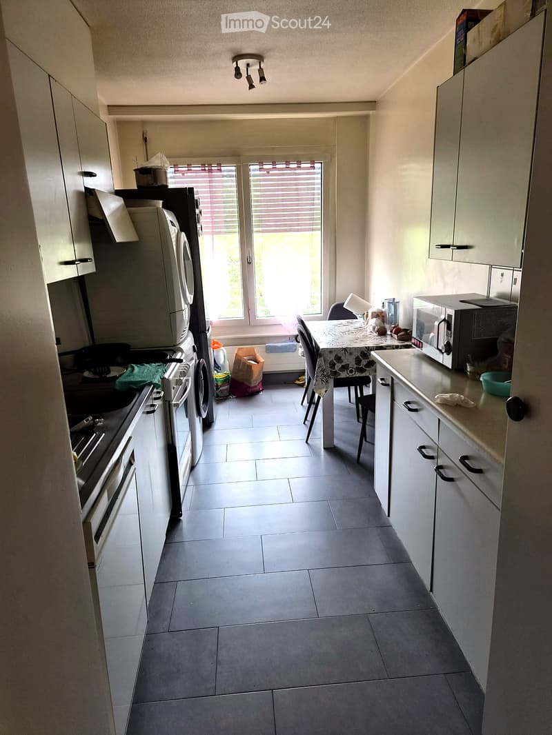 Modern kitchen with white cabinets, tiled floor, and stainless steel appliances including a refrigerator, oven, and microwave. A small dining area with a table and chairs is visible near the window.