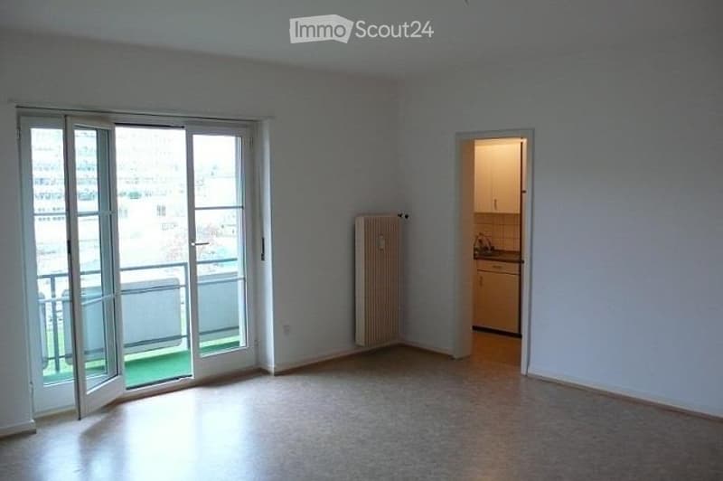 Empty living space with large sliding glass doors leading to a balcony, a radiator, and an adjacent small kitchen area.