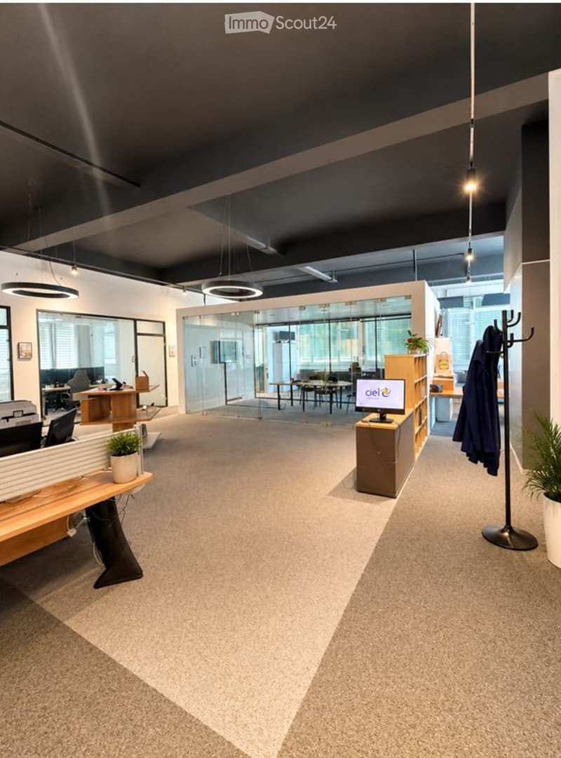 Modern office space with a reception area, desks, chairs, and a coat rack. The room features a sleek design with glass partitions and pendant lighting.