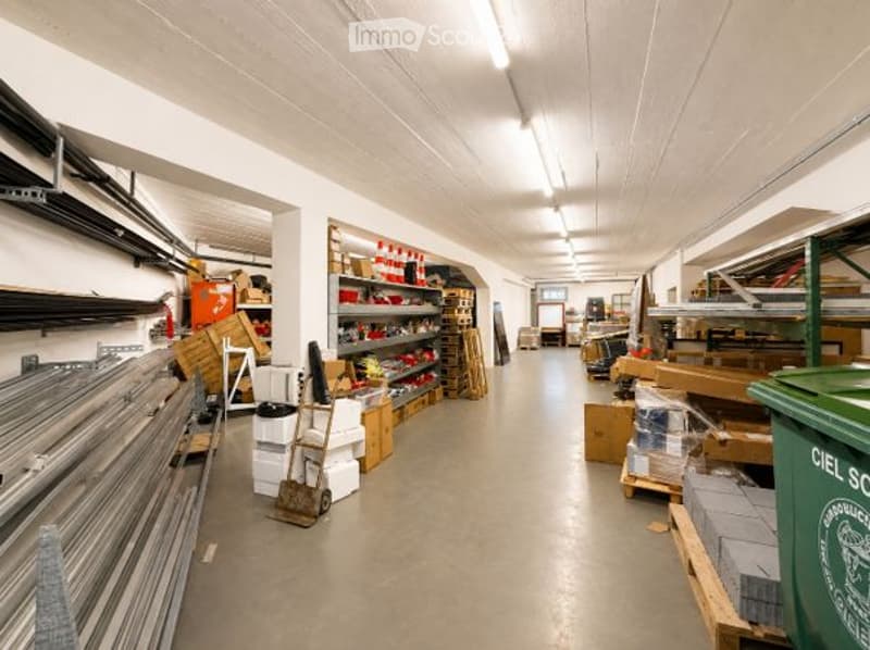 A spacious, well-lit warehouse or storage area with exposed white ceiling beams, metal shelving units, and various construction materials and tools. The floor is concrete, and the space is organized with pallets, boxes, and stacked items.