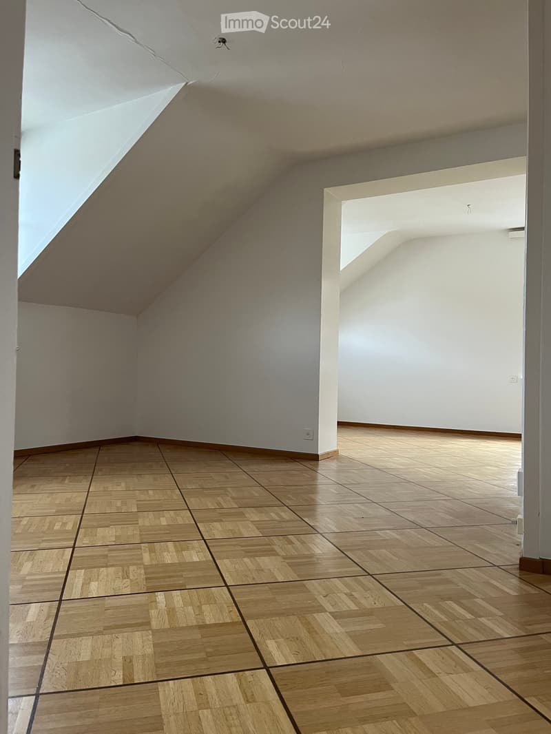 Empty room with wooden parquet flooring and white walls, featuring a slanted ceiling and visible electrical outlets.