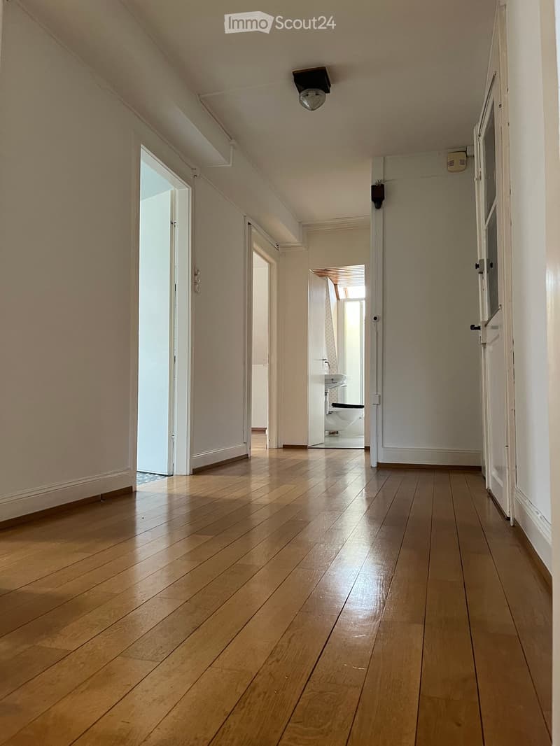A narrow hallway with wooden flooring, white walls, and several doors leading to different rooms, including a visible bathroom with a sink and toilet.