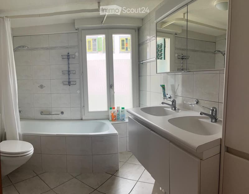 Bathroom with a bathtub, toilet, double sink, mirror, and various toiletries on the sink counter. The room features tiled walls and a window with shutters.