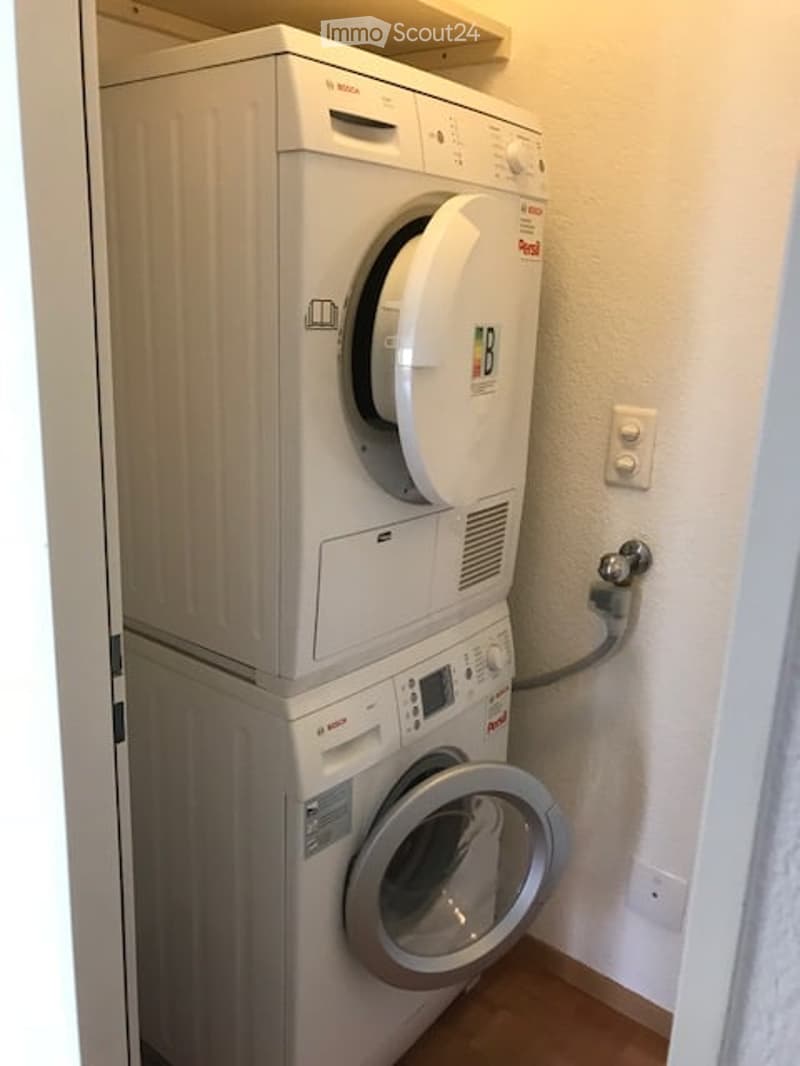 Compact bathroom with stacked Bosch washing machine and dryer unit, white tiled walls, wooden floor, and light switch.