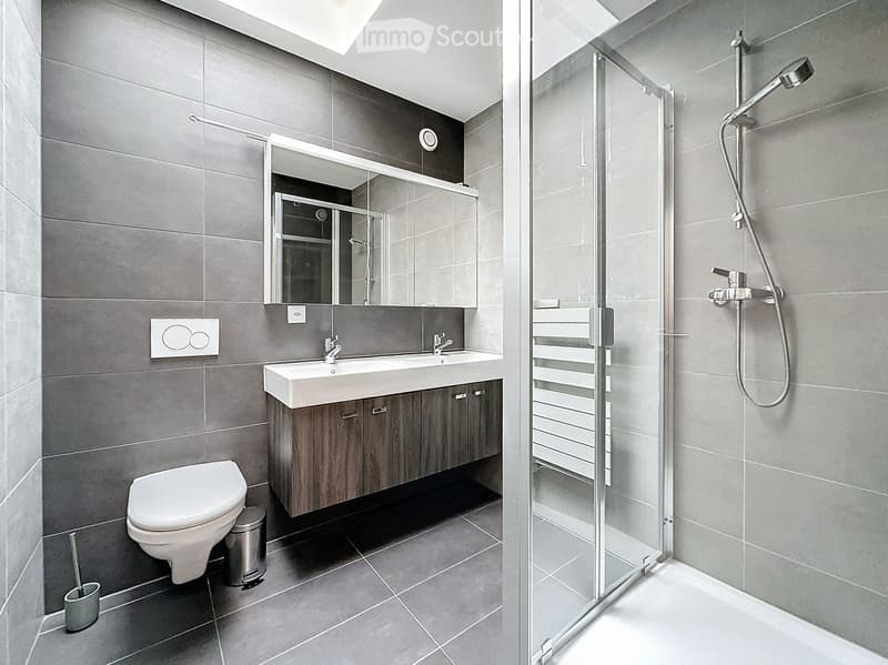 Modern bathroom with grey tiles, a double sink vanity with wooden cabinets, a toilet, a shower with glass enclosure, and a large mirror above the sink.