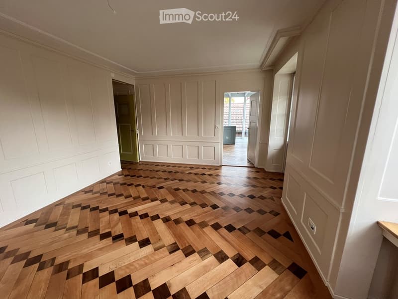 Empty hallway with wooden herringbone flooring and white paneled walls, featuring several doors and an open doorway leading to another room.