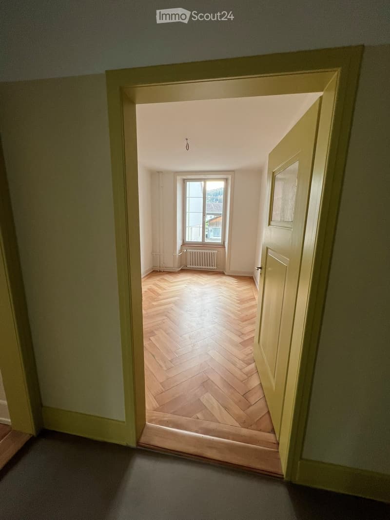 Empty room with wooden herringbone flooring, white walls, a window with a radiator below, and a partially open door leading to another room.