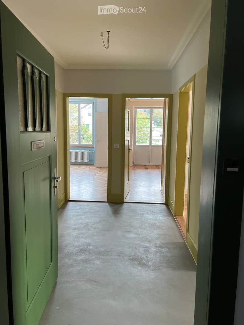 Empty hallway with grey concrete floor, light green walls, and two open doors leading to rooms with wooden flooring and large windows.