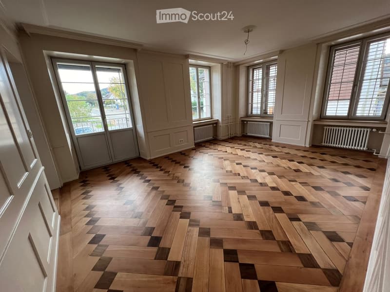 Empty room with wooden herringbone flooring, large windows, radiators, and white walls.