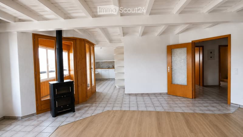 Bright, open living space with wooden floors, white walls, and a black wood-burning stove. Features include wooden doors, built-in wooden shelves, and a small kitchen area with white cabinets.
