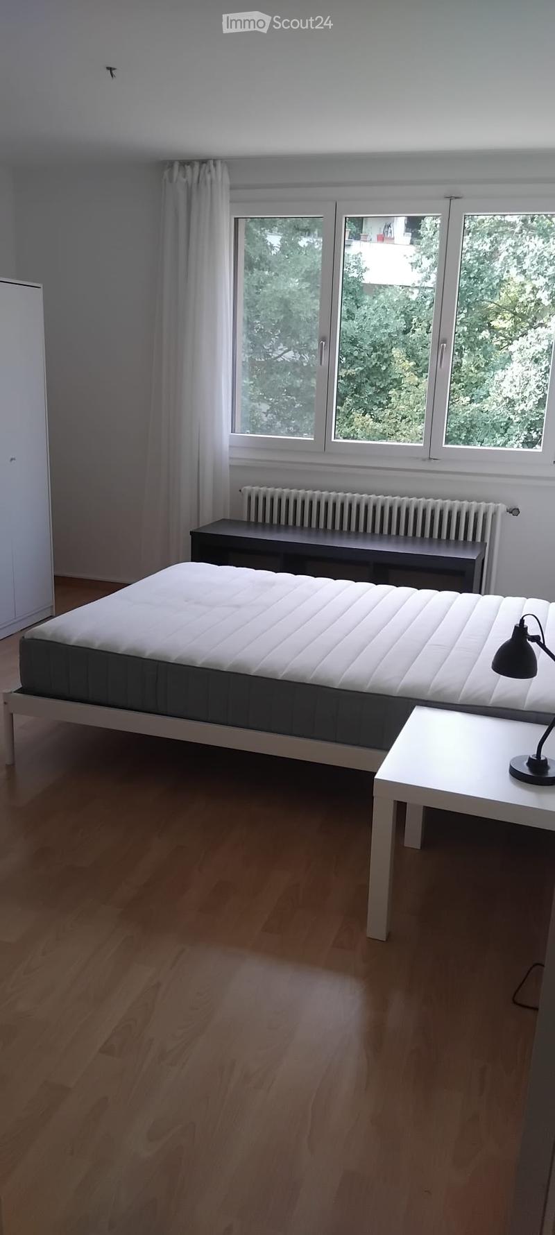 Minimalist bedroom with a white bed, small white table with a black lamp, bench, radiator, and large windows with white curtains.