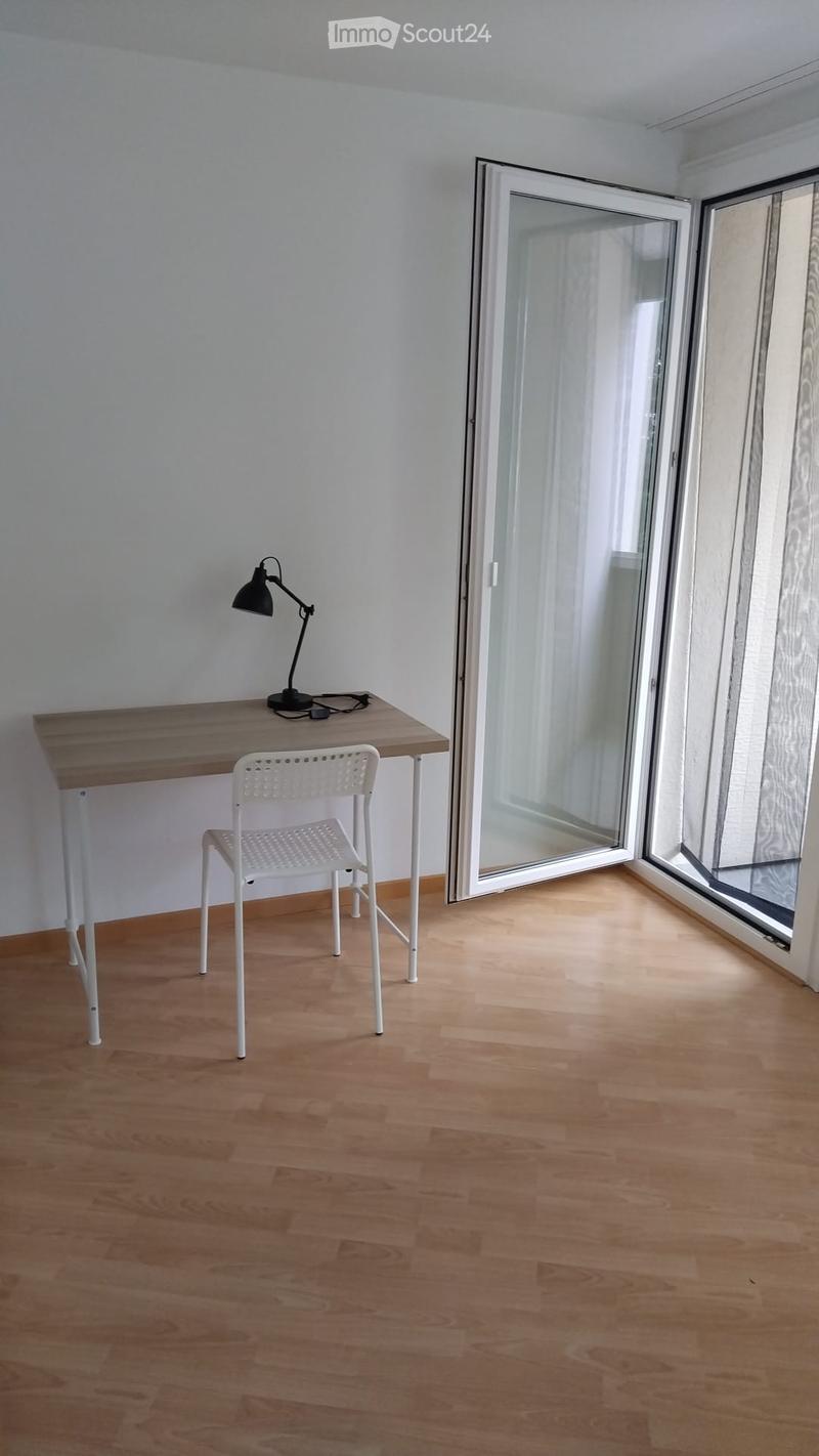 A small, modern room with light wooden flooring, a white desk, a white chair, and a black desk lamp. Large glass doors lead to a balcony.