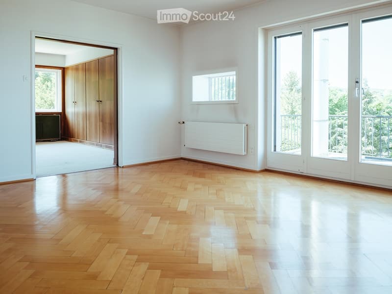 Empty room with wooden herringbone flooring, white walls, large windows, and sliding glass doors leading to a balcony.