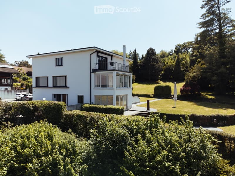 A two-story white house with large windows and a balcony, surrounded by a well-maintained garden and trees.