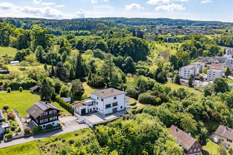 Aerial view of a suburban neighborhood with multiple houses, lush greenery, and a scenic backdrop of hills and forests.