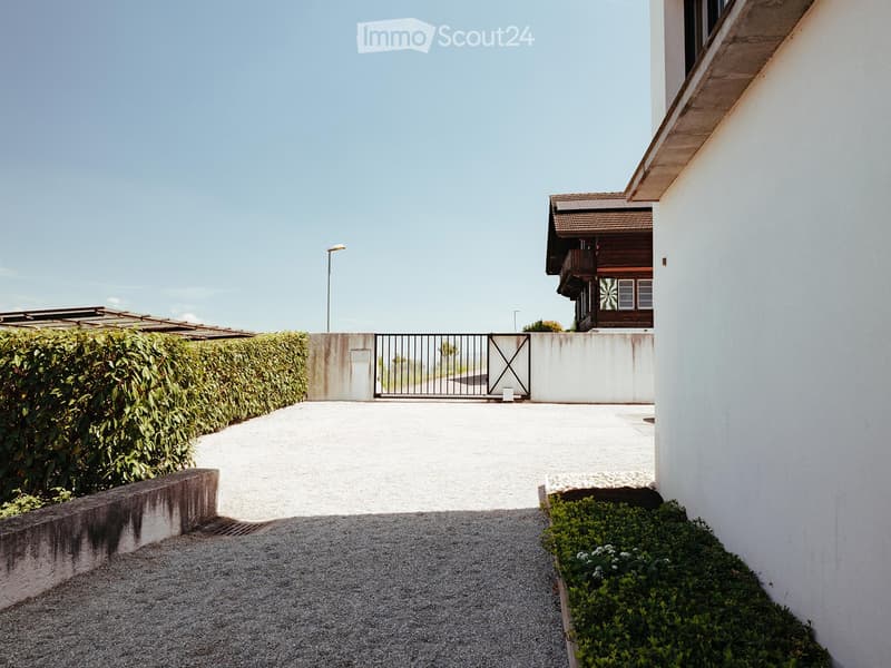 A gated driveway leads to a house with a balcony and a wooden structure, surrounded by neatly trimmed hedges and small plants.